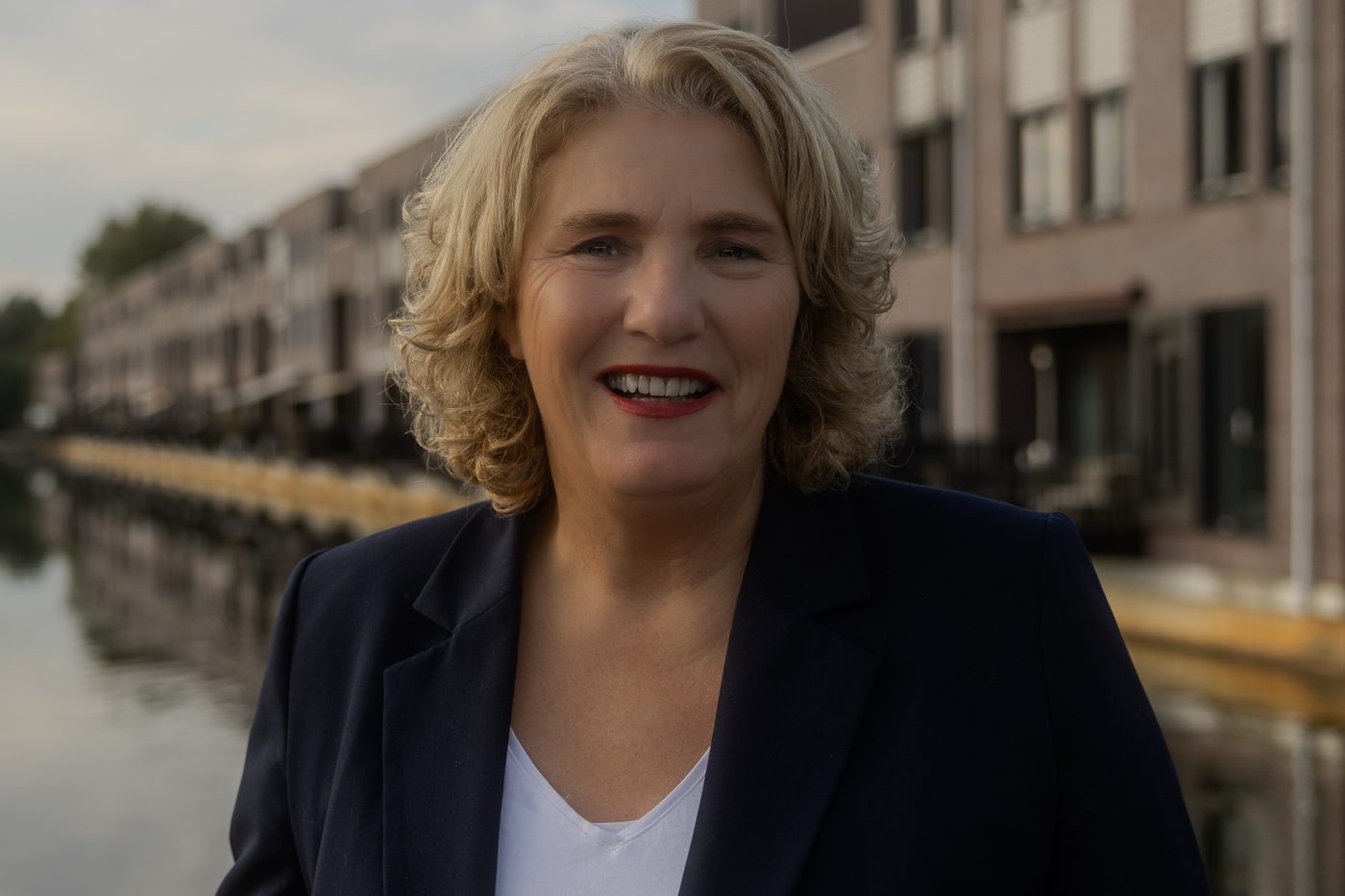 Makelaar in donker colbert lachend, met grachtpanden en water achter haar